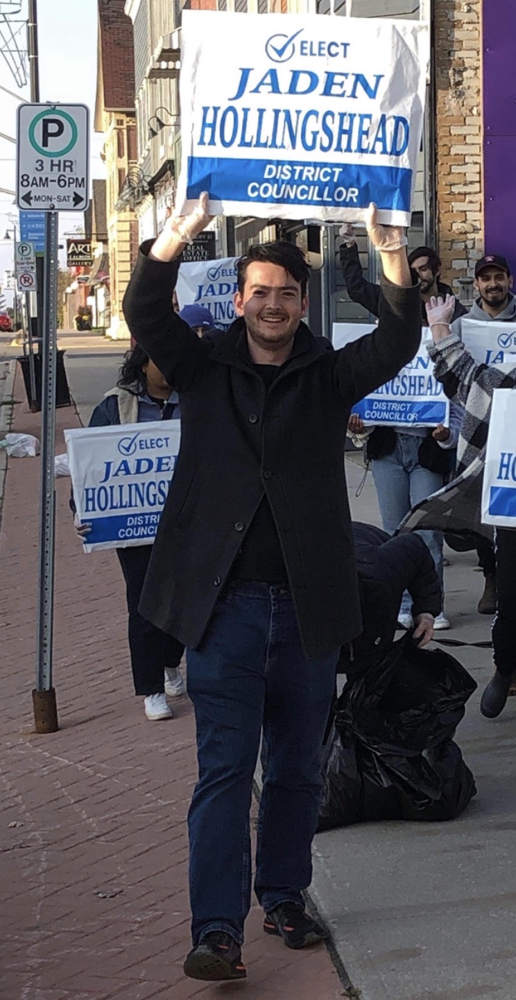 Jaden Hollingshead holding an ELECT JADEN HOLLINGSHEAD &mdash; DISTRICT COUNCILLOR sign above his head on a downtown sidewalk, with volunteers carrying signs behind him.